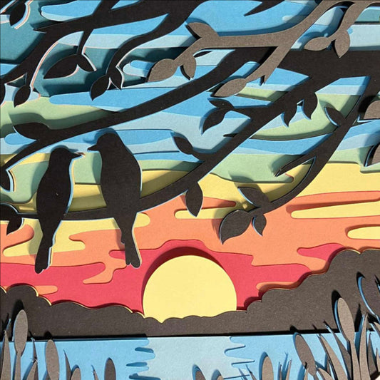 A colorful layered paper-cut style artwork showing two black birds perched on branches over a sunset landscape with blue sky, yellow sun, orange and red sky layers, dark land, and water reeds, presented on a black background.
