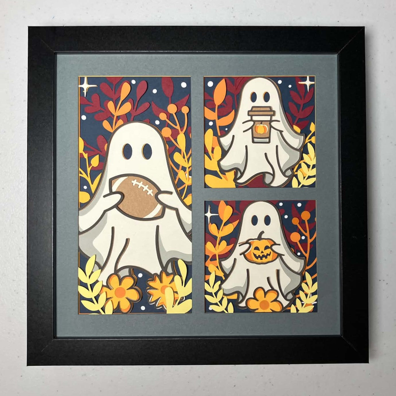 Framed three-panel Halloween art print featuring cartoon ghosts in autumn colors, shown against a light gray wall. The left panel shows a ghost holding a football, the top-right shows a ghost holding a coffee cup, and the bottom-right shows a ghost holding a jack-o'-lantern.
